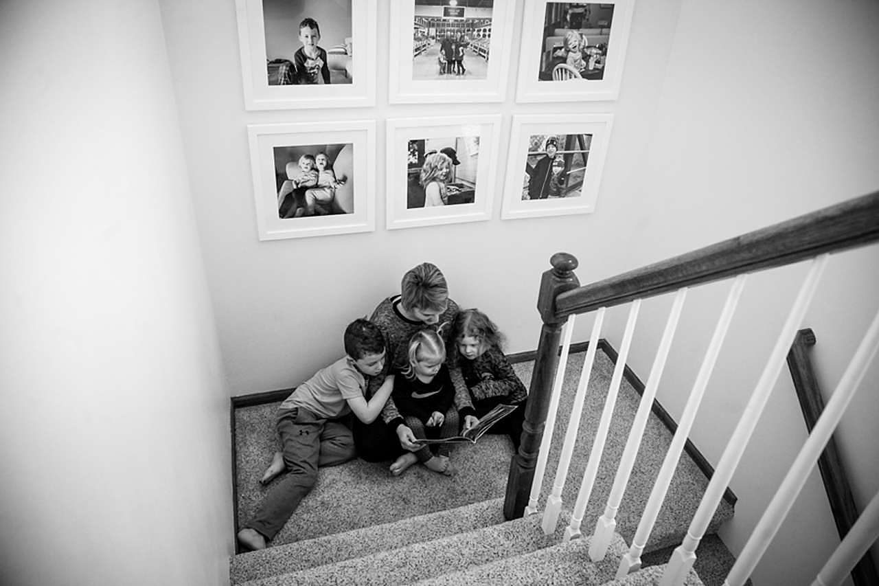 Mom reading book to kids at bottom of stairs under framed family photos